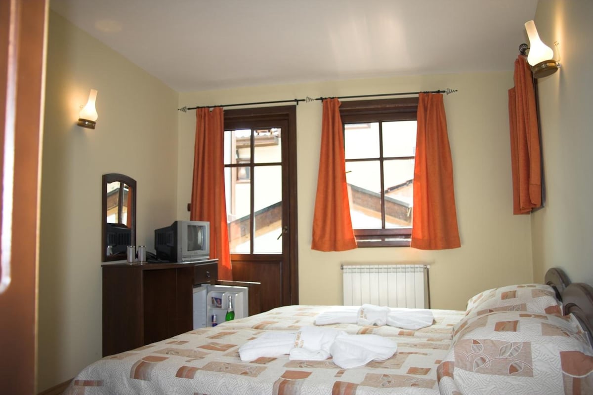 A hotel room with a bed, a television on a wooden dresser, and a window with orange curtains.