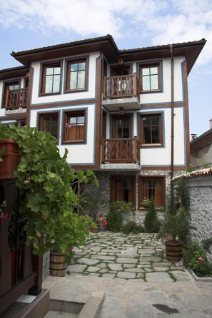 Un edificio de dos plantas con fachada de piedra, varias ventanas con contraventanas de madera y un balcón central con barandilla decorativa.