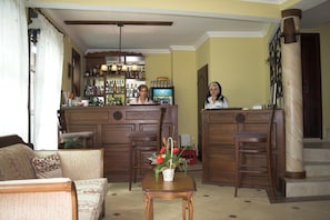 A bar area with a counter, shelves of bottles, and a television screen. There is a reception desk with a person seated behind it. - Hotel Old Times (Asenovgrad)