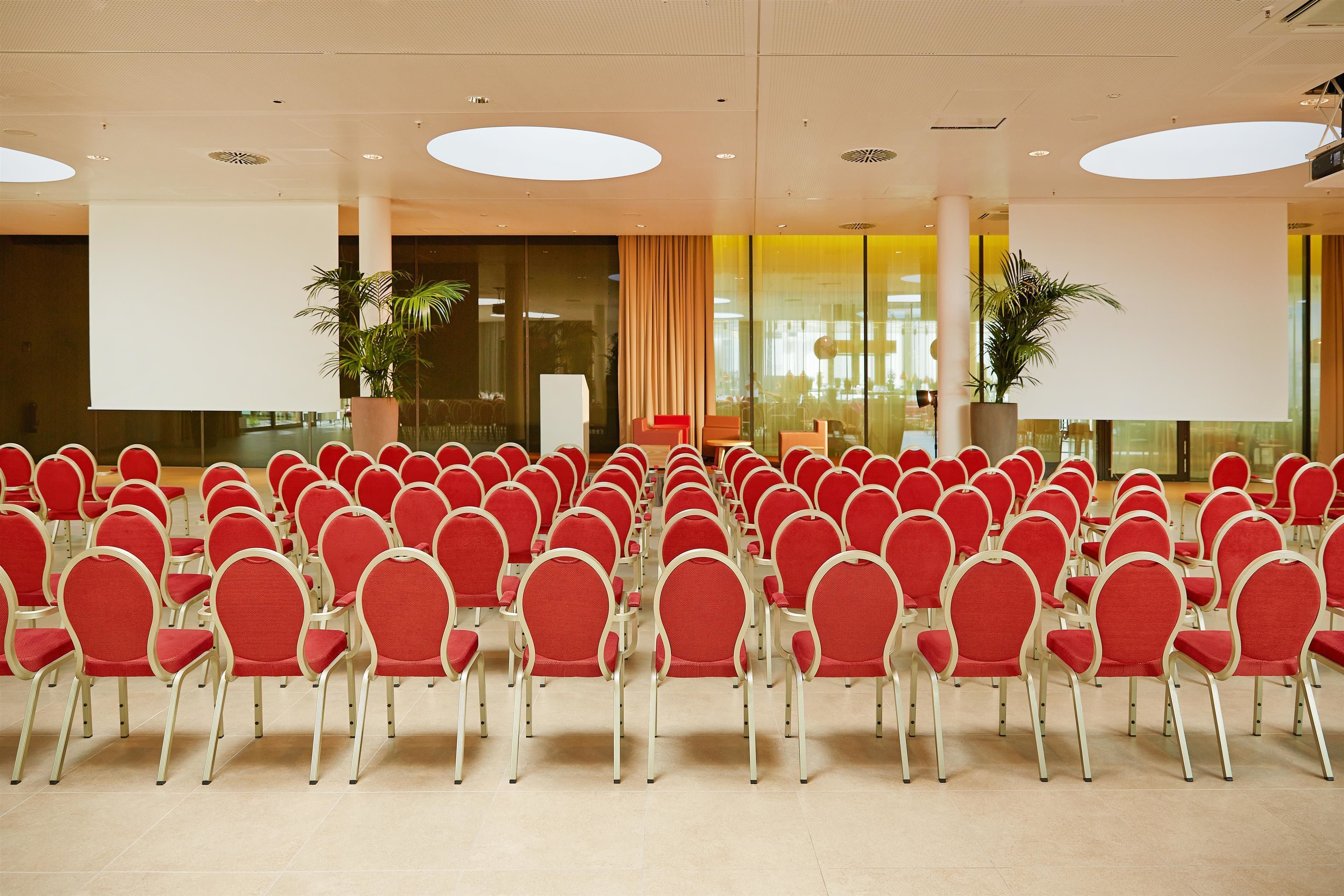 Une salle de conférence avec des rangées de chaises rouges, de grandes fenêtres et des plantes en pot.