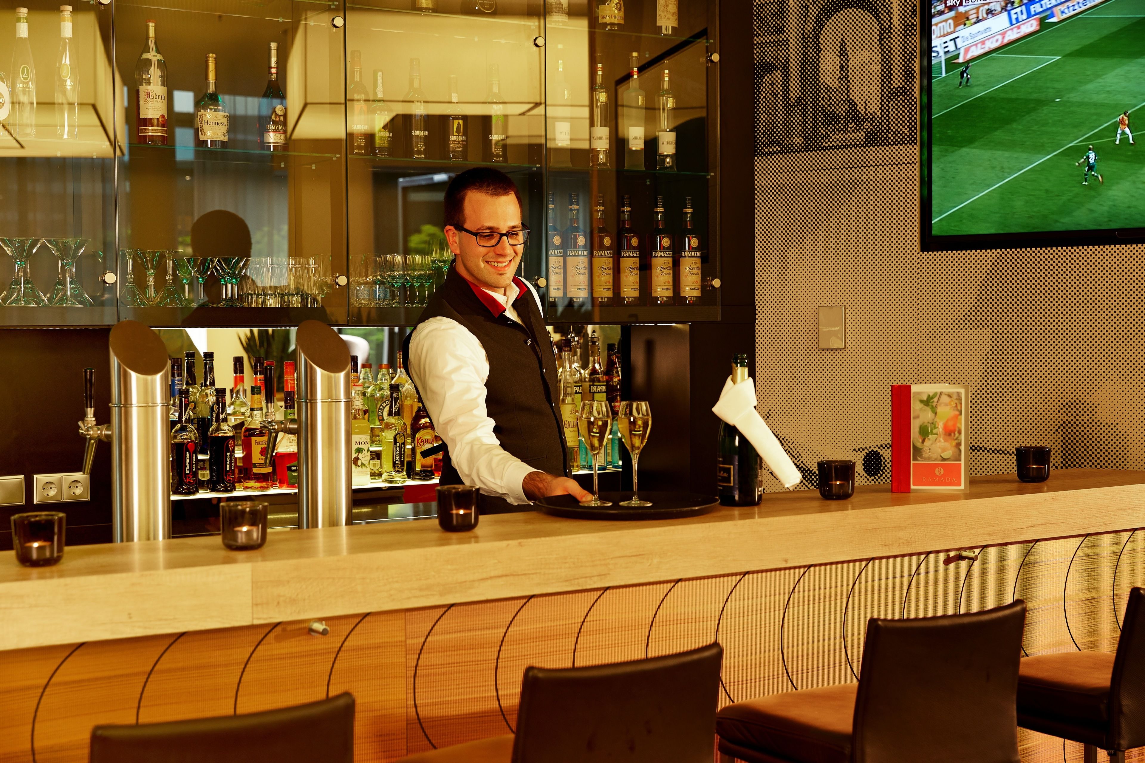 A bar area with a bartender, shelves of liquor bottles, glasses, and a television displaying a soccer game.