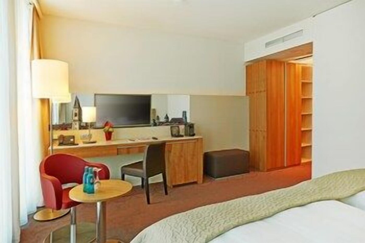 A hotel room with a bed, a desk, a chair, a small table, a TV, and a wardrobe.