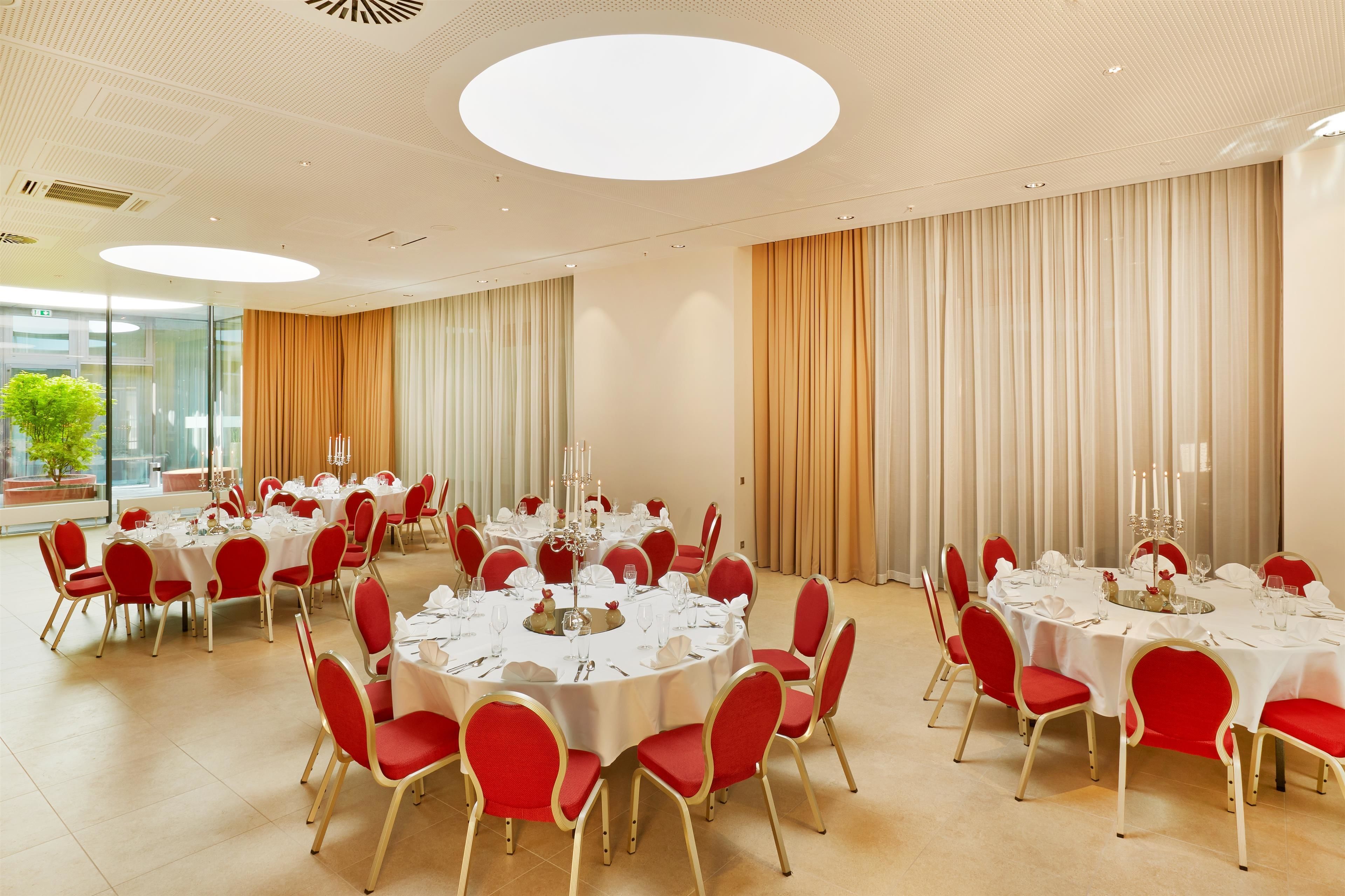 A banquet hall with multiple round tables set for dining, red upholstered chairs, and a large window providing natural light.