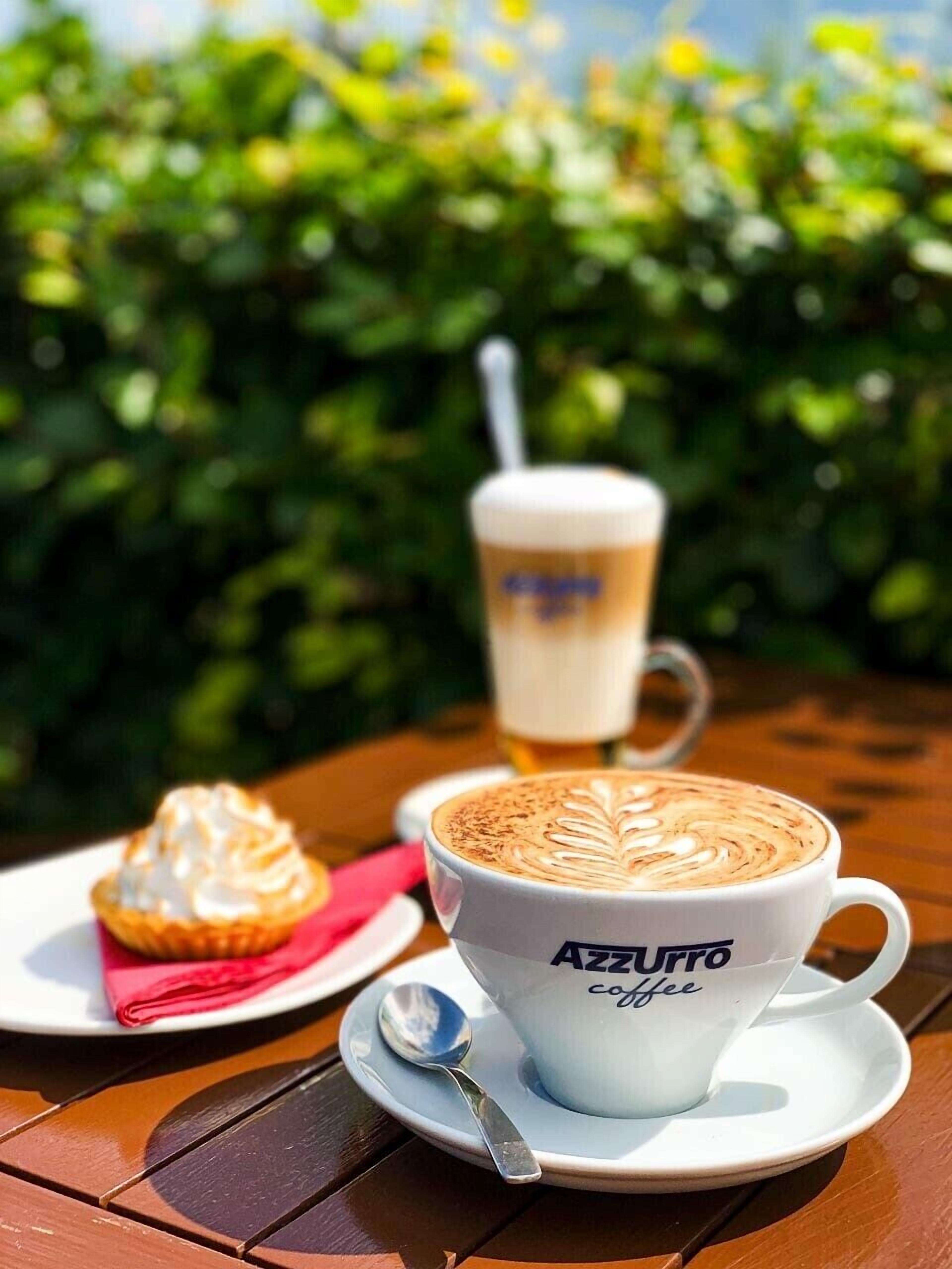 Een kop koffie met latte-art op een schotel, een muffin op een bord, en een glas bier met een rietje op een tafel.