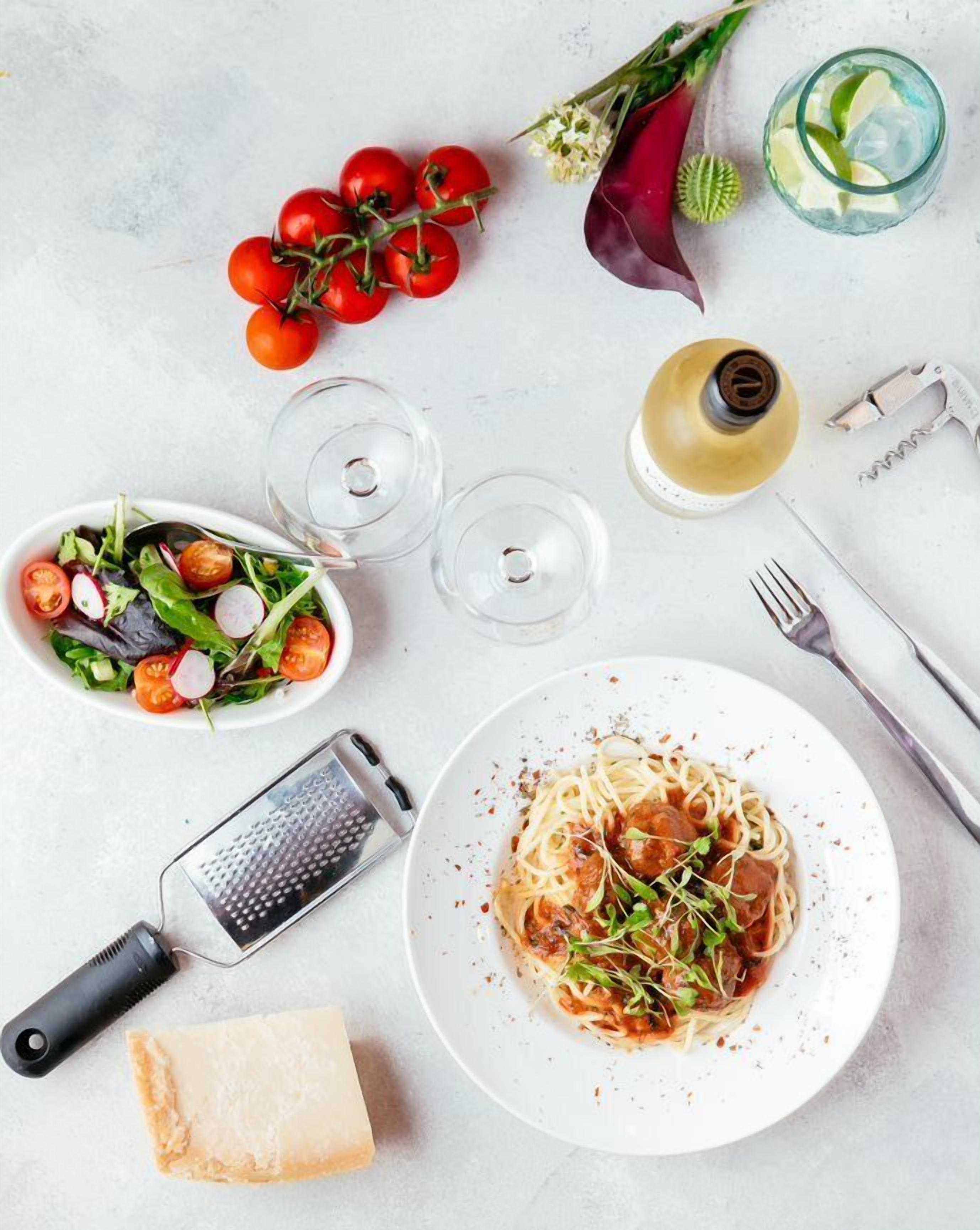 Une table à manger avec une assiette de spaghettis, un saladier de salade, un morceau de fromage, une râpe et une bouteille d’huile d’olive.