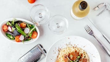 Una mesa de comedor con un plato de espagueti, un bol de ensalada, un bloque de queso, un rallador y una botella de aceite de oliva.
