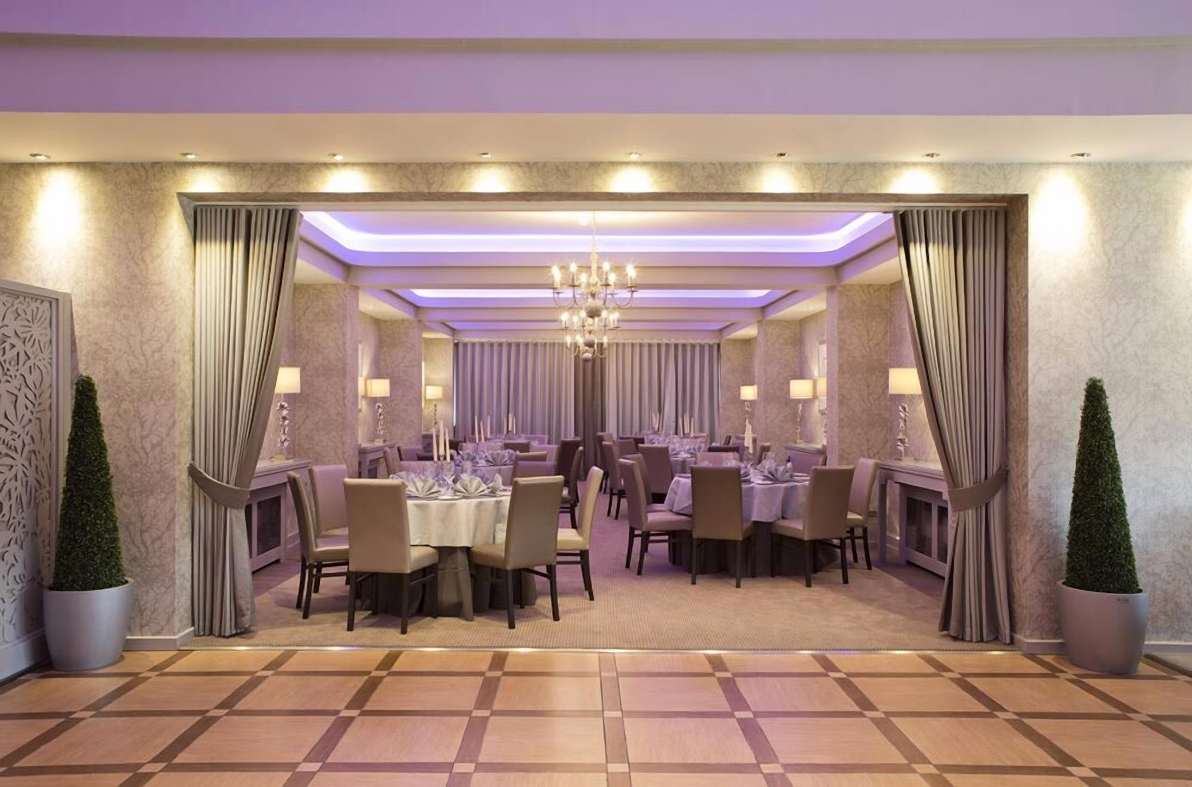 A formal dining room with tables set for guests, a chandelier, and decorative curtains.