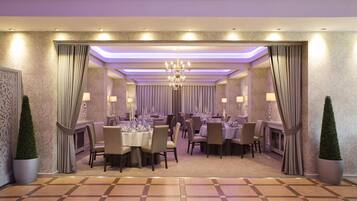 A formal dining room with tables set for guests, a chandelier, and decorative curtains.