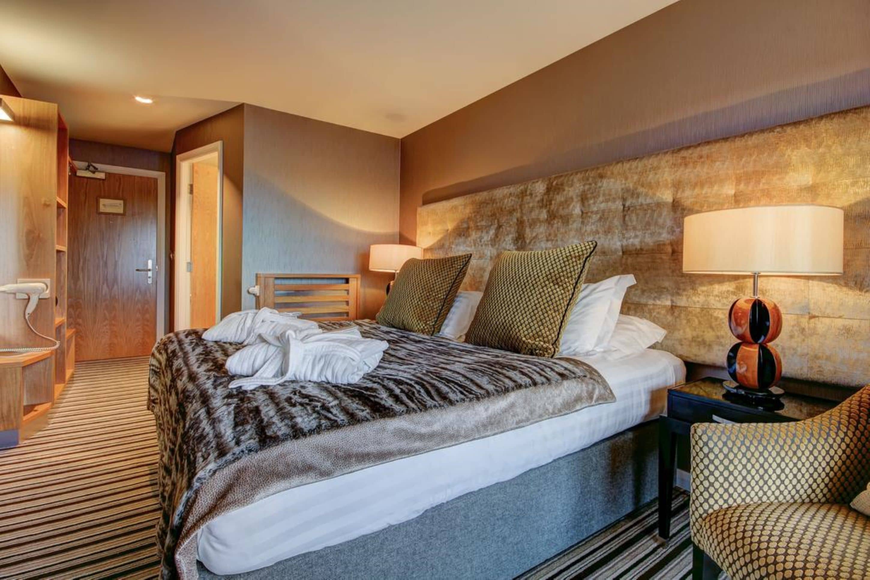 A hotel room with a large bed, a wooden chair, a nightstand with a lamp, and a wall-mounted headboard.
