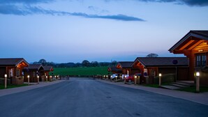A row of wooden cabins with a paved road leading to a gate, set against a twilight sky. - Ingliston Country Club Hotel (Bishopton)