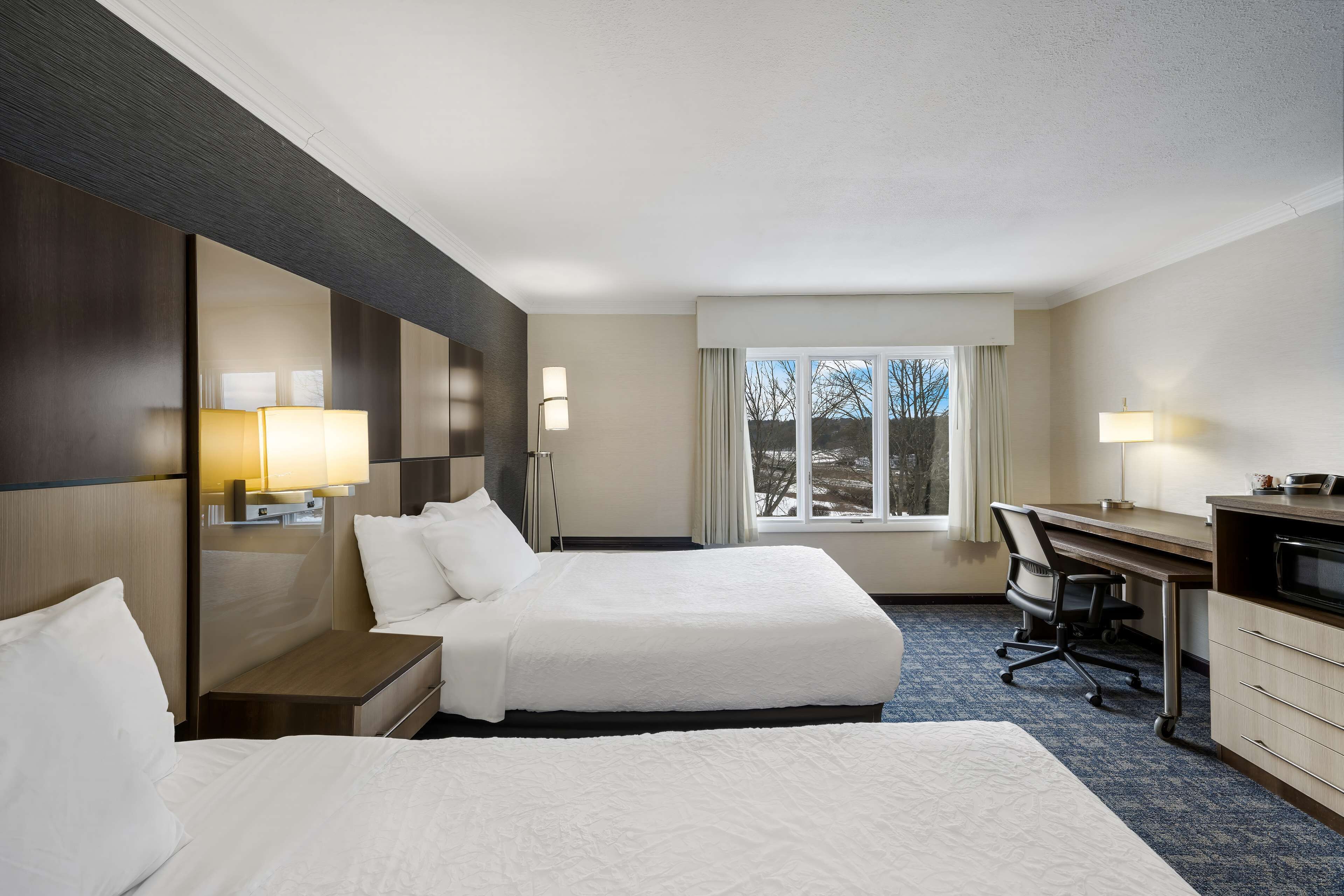 Quarto de hotel com duas camas, uma escrivaninha, uma cadeira, uma televisão e uma janela com vista para árvores.