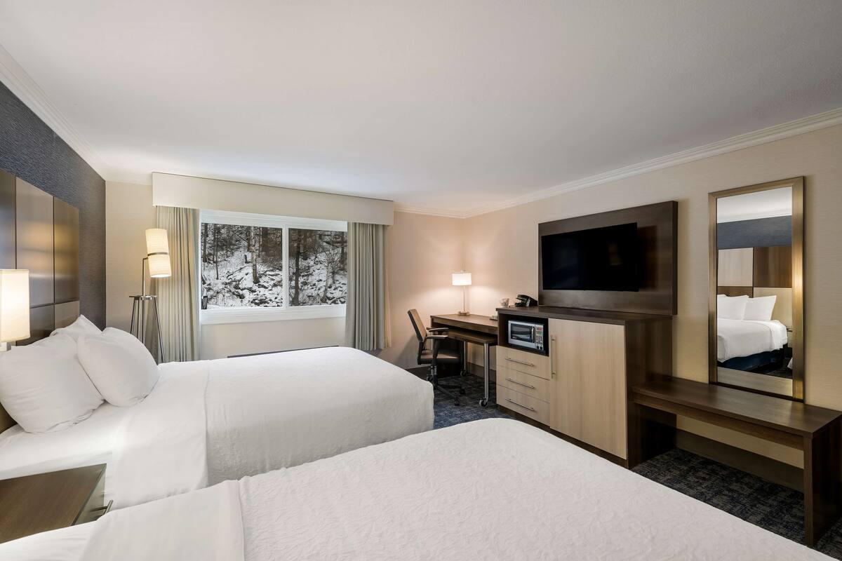 A hotel room with two beds, a desk with a chair, a TV, a mirror, and a window with a view of trees.