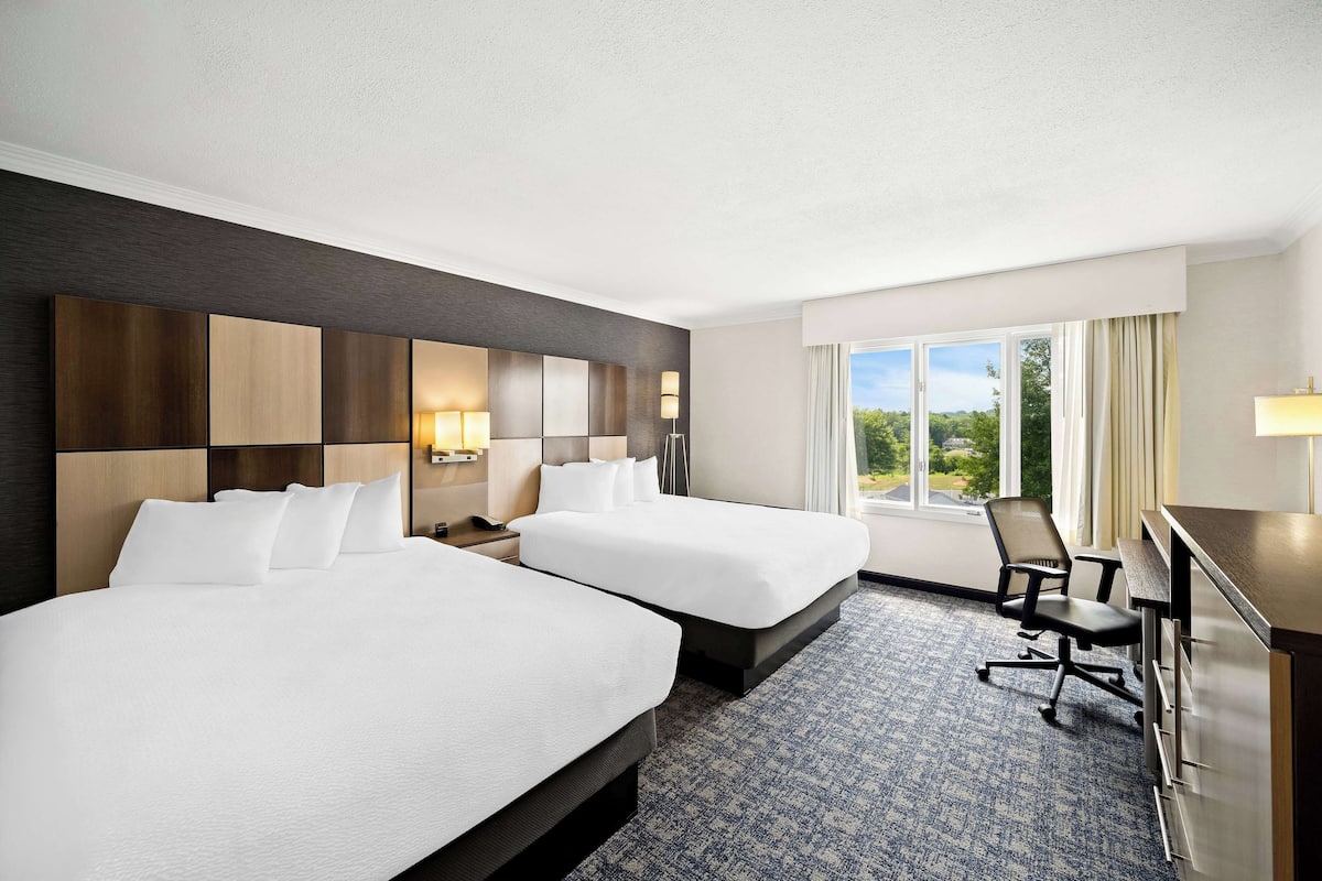 A hotel room with two beds, a desk, and a view of the outdoors.