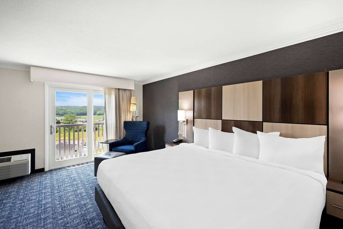 A hotel room with a large bed, a blue armchair, a nightstand, and a view of a balcony with a green landscape.