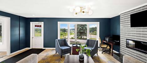 A living room with two armchairs, a coffee table, and a TV. There is a door leading outside, a window with a view, and a patterned wall.