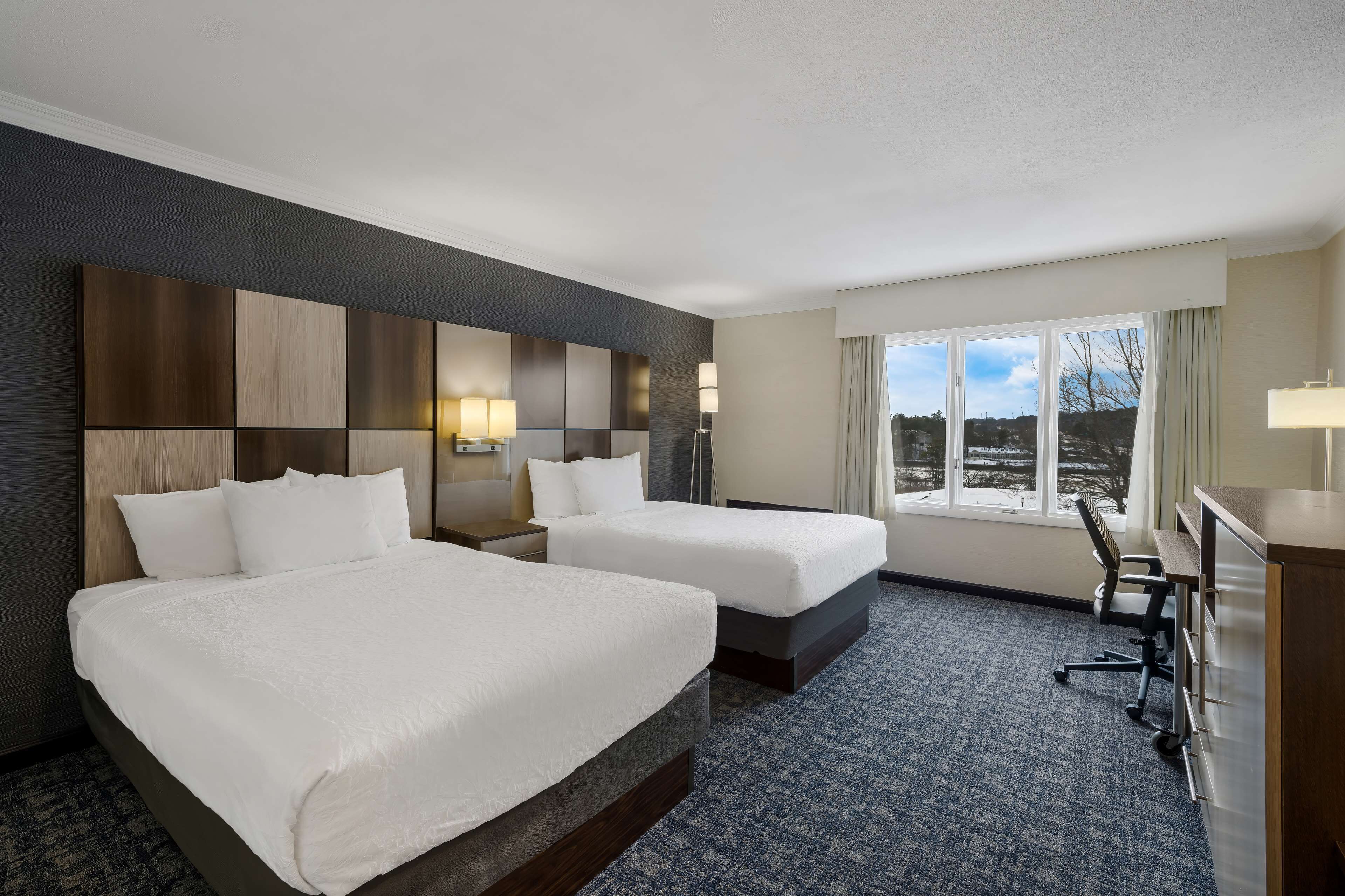A hotel room with two beds, a desk, and a view of the outdoors.