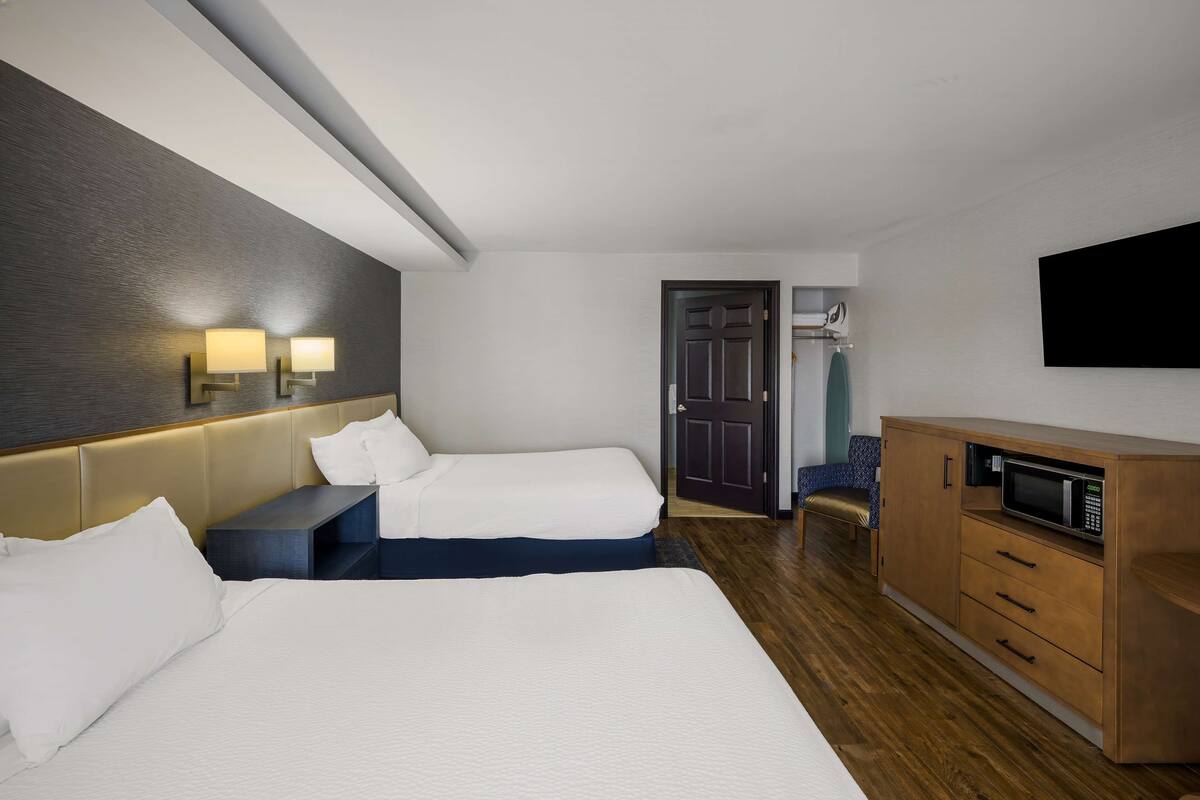 A hotel room with two beds, a wooden cabinet with a microwave, a flat-screen TV, and a door leading to a bathroom.