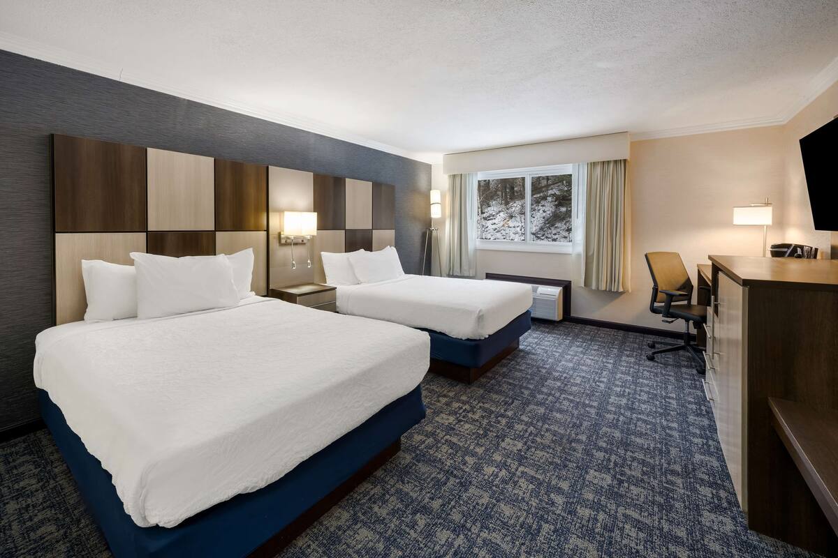 A hotel room with two beds, a desk, a chair, a TV, and a window with a view of snow outside.