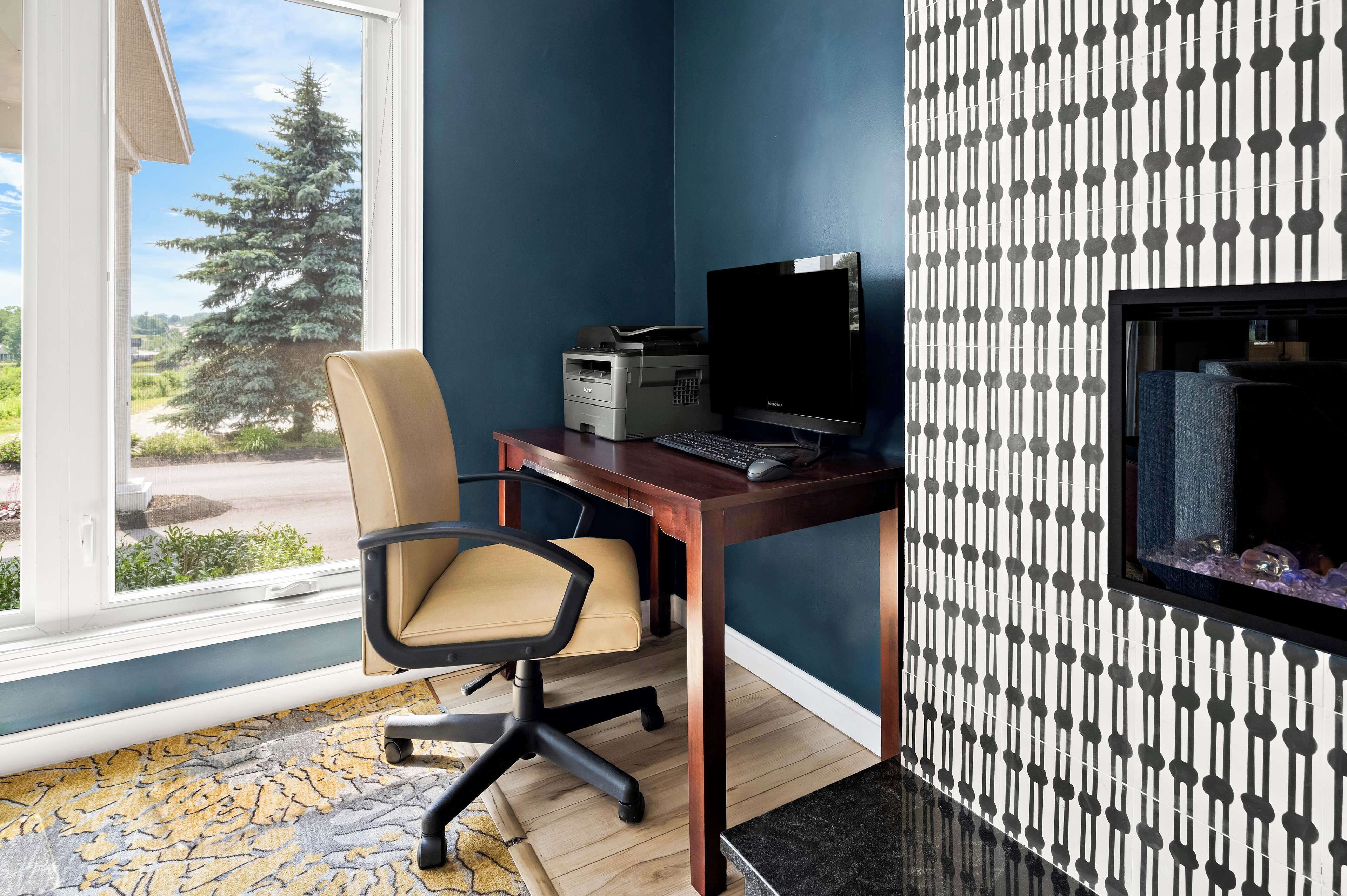 A room with a desk, chair, and a computer. There is a window with a view of trees and a building.
