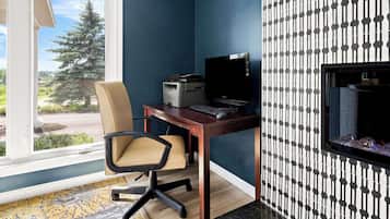 A room with a desk, chair, and a computer. There is a window with a view of trees and a building.