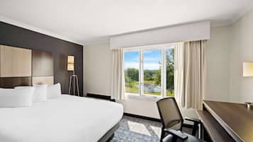 Habitación de hotel con una cama grande, un escritorio, una silla, una lámpara y una ventana con vistas a zonas verdes.