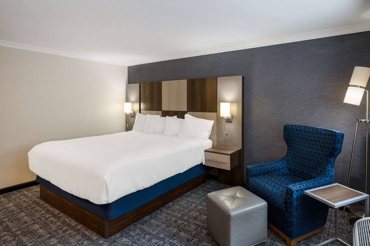 A hotel room with a large bed, a nightstand, a blue patterned armchair, a small table, and a lamp.