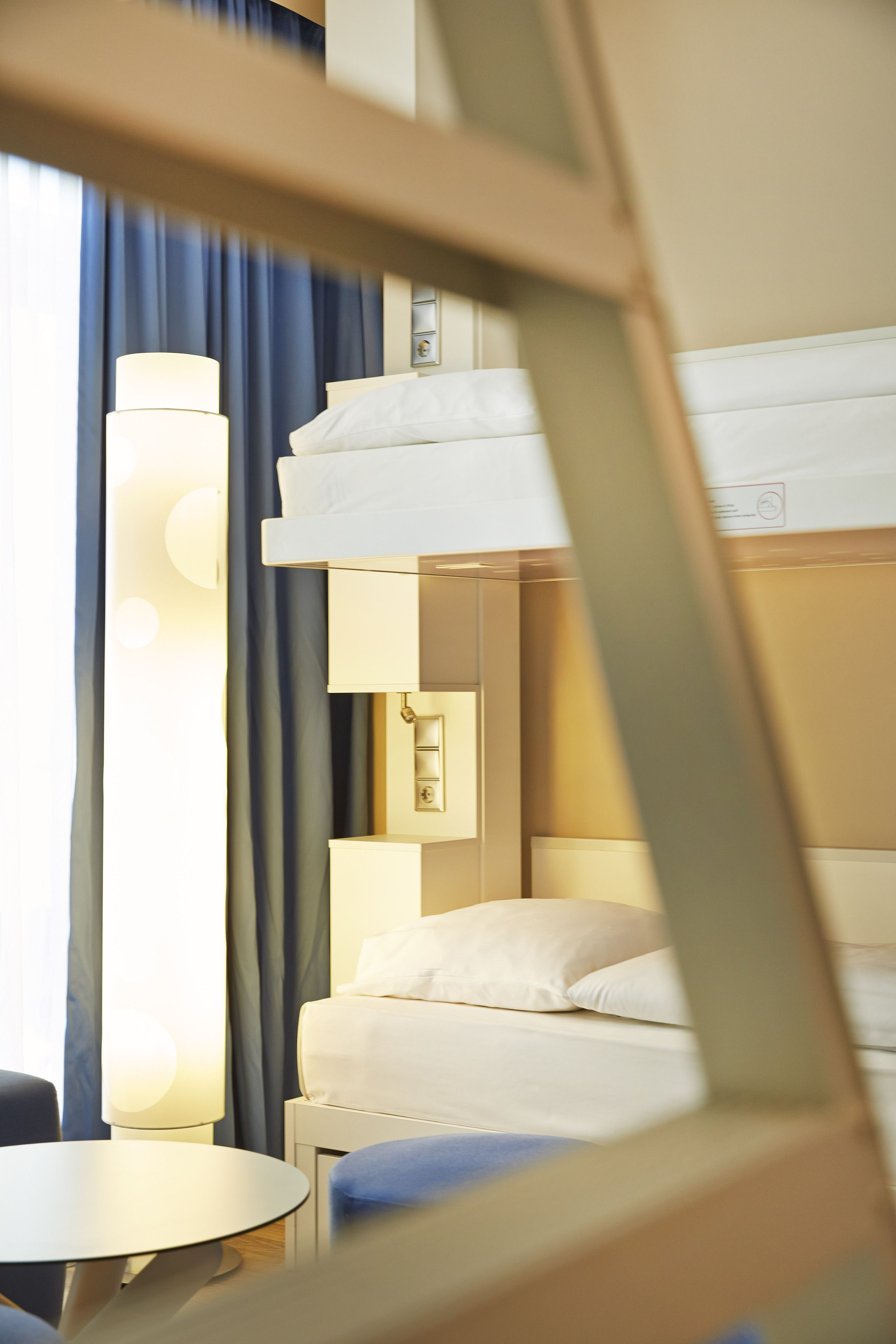 A hotel room with a bunk bed, a bedside table, a lamp, and a window with curtains.