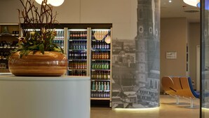 A modern interior with a large digital screen displaying a silhouette, a vending machine with beverages, and a seating area with blue chairs. - H2 Hotel München Messe (Munich)