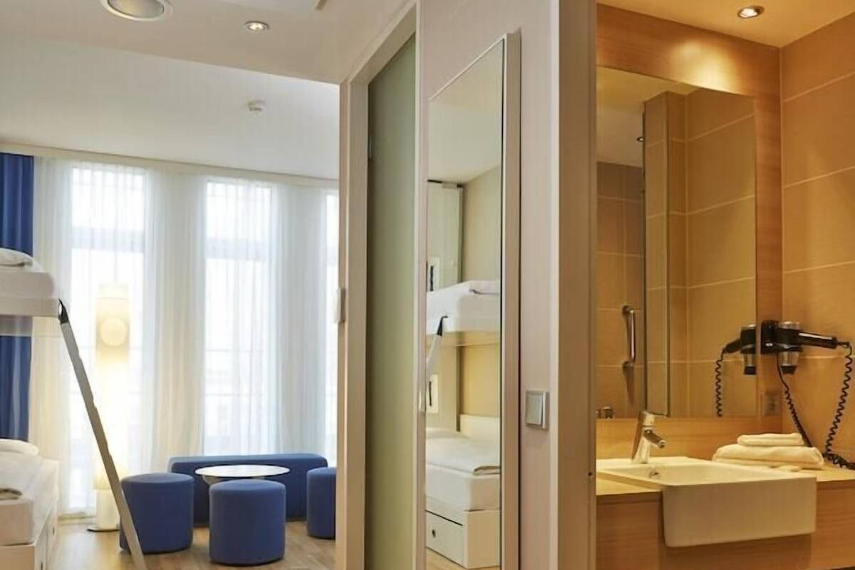 Una habitación de hotel moderna con piso de madera, un sofá, otomanas, un espejo, un lavamanos y un secador de pelo.