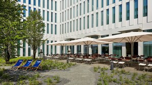 An outdoor seating area with tables and chairs, a pathway, and a building with large windows in the background. - H2 Hotel München Messe (Munich)