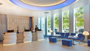 A modern hotel reception area with a large H2 Hotels sign, a reception desk, seating area, and large windows offering a view of the outdoors. - H2 Hotel München Messe (Munich)