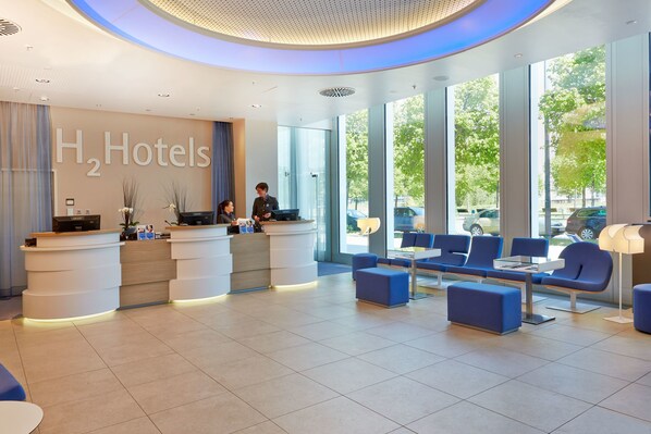 A modern hotel reception area with a large H2 Hotels sign, a reception desk, seating area, and large windows offering a view of the outdoors. - H2 Hotel München Messe (Munich)