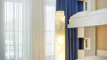 A hotel room with a bunk bed, a round table, two ottomans, and a floor lamp.