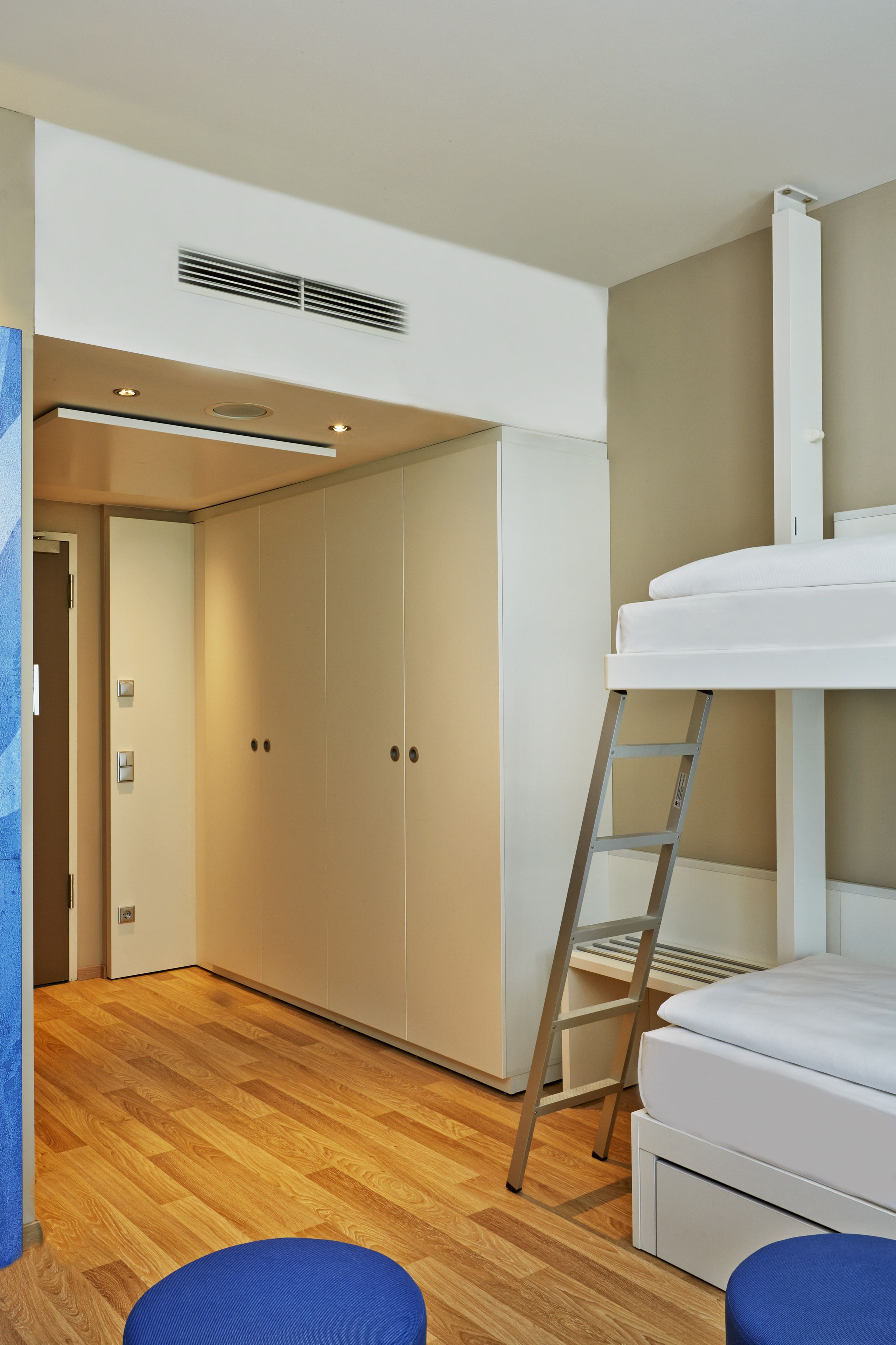 A bunk bed with a ladder, a wardrobe, and a small stool in a room with wooden flooring and a door.