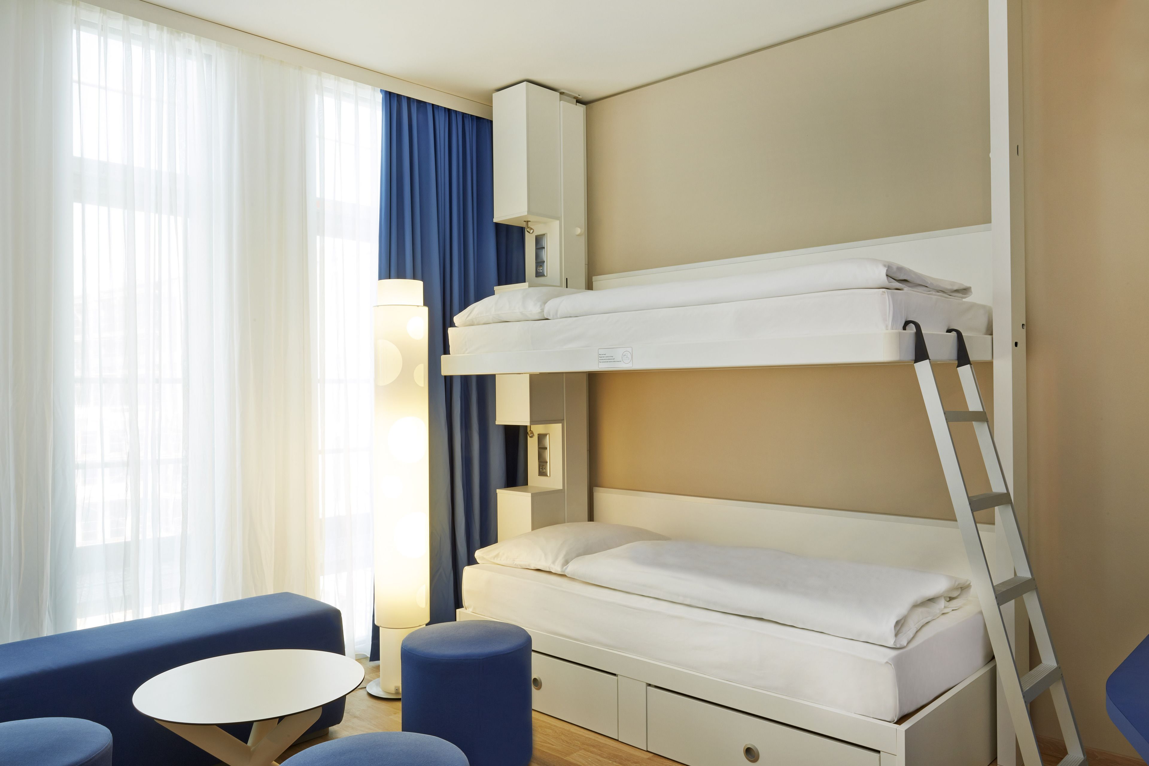 A bunk bed with a ladder, a small table, and a blue chair in a room with a window and curtains.