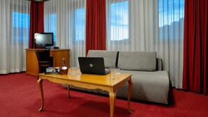 A hotel room with a sofa, a wooden table with a laptop and a bottle, a TV, and red curtains. - Real Hotel Veliko Tarnovo (Veliko Tarnovo)