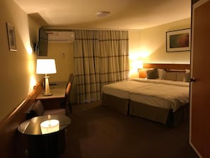 A hotel room with a bed, a desk with a lamp, a chair, a round table with a lamp, and a window with curtains.