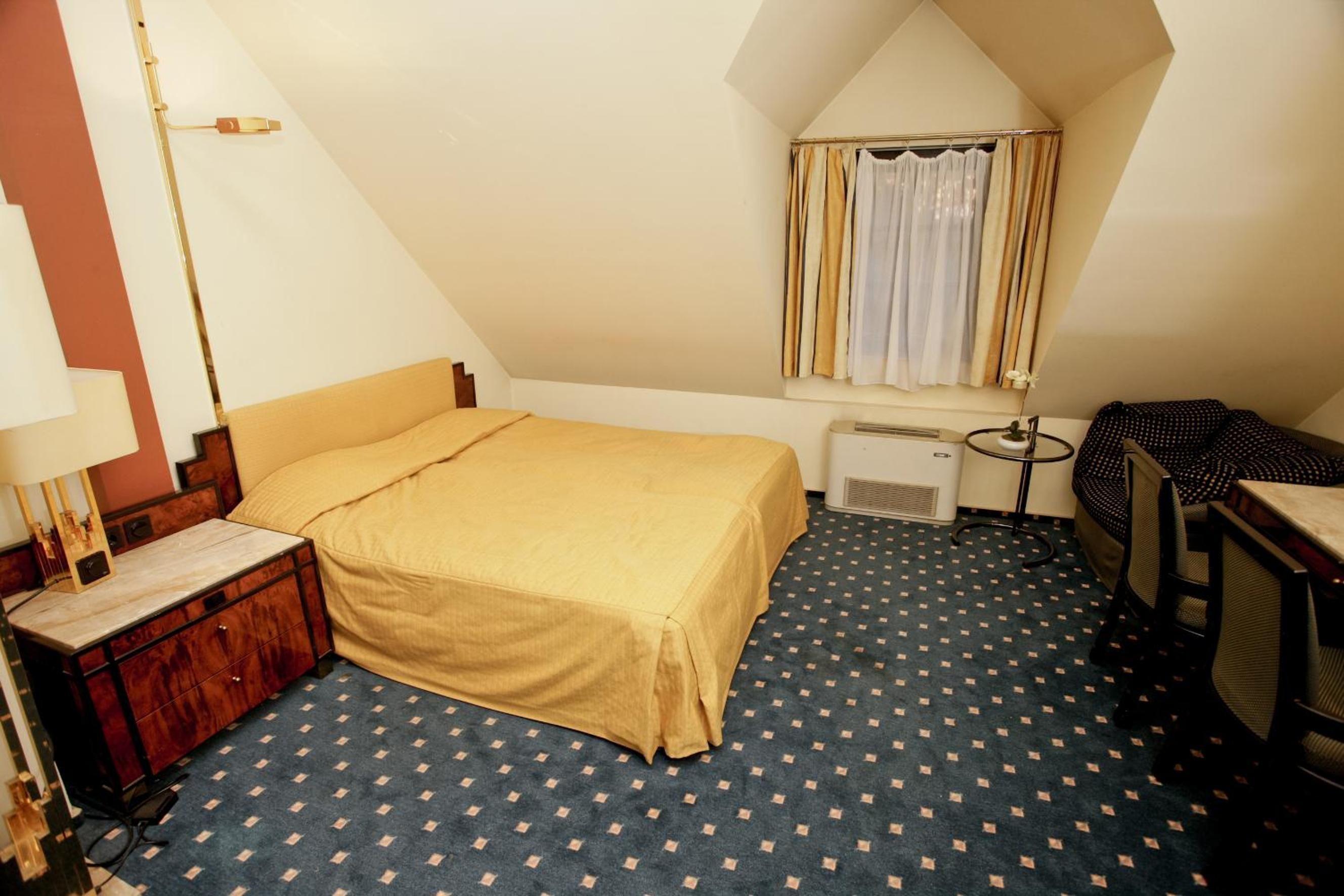 Une petite pièce triangulaire comprenant un lit, un bureau et une fenêtre avec des rideaux.