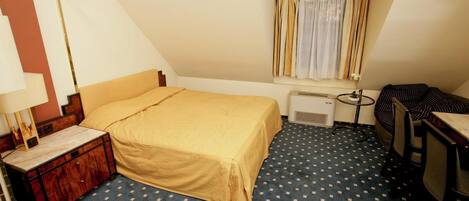 Une petite pièce triangulaire comprenant un lit, un bureau et une fenêtre avec des rideaux.