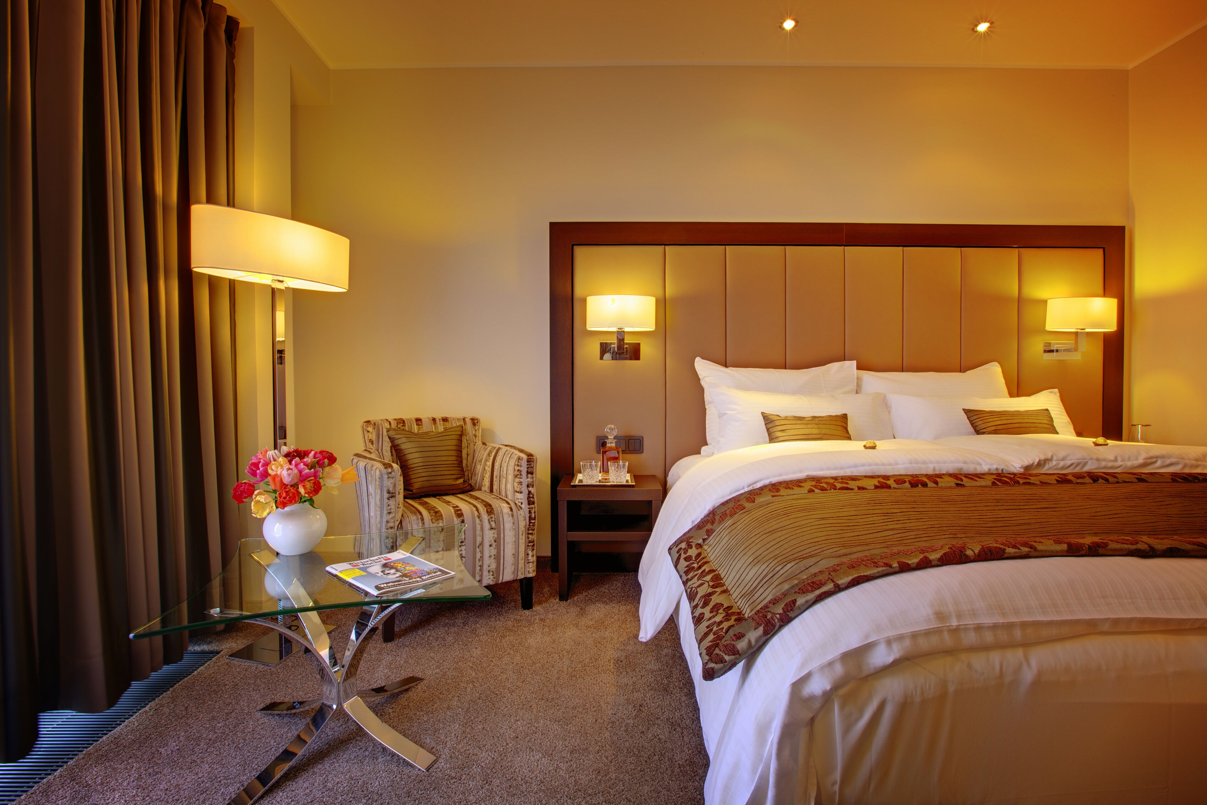 Habitación de hotel con una cama grande, una mesa de vidrio con un jarrón de flores, una silla y dos lámparas de noche.