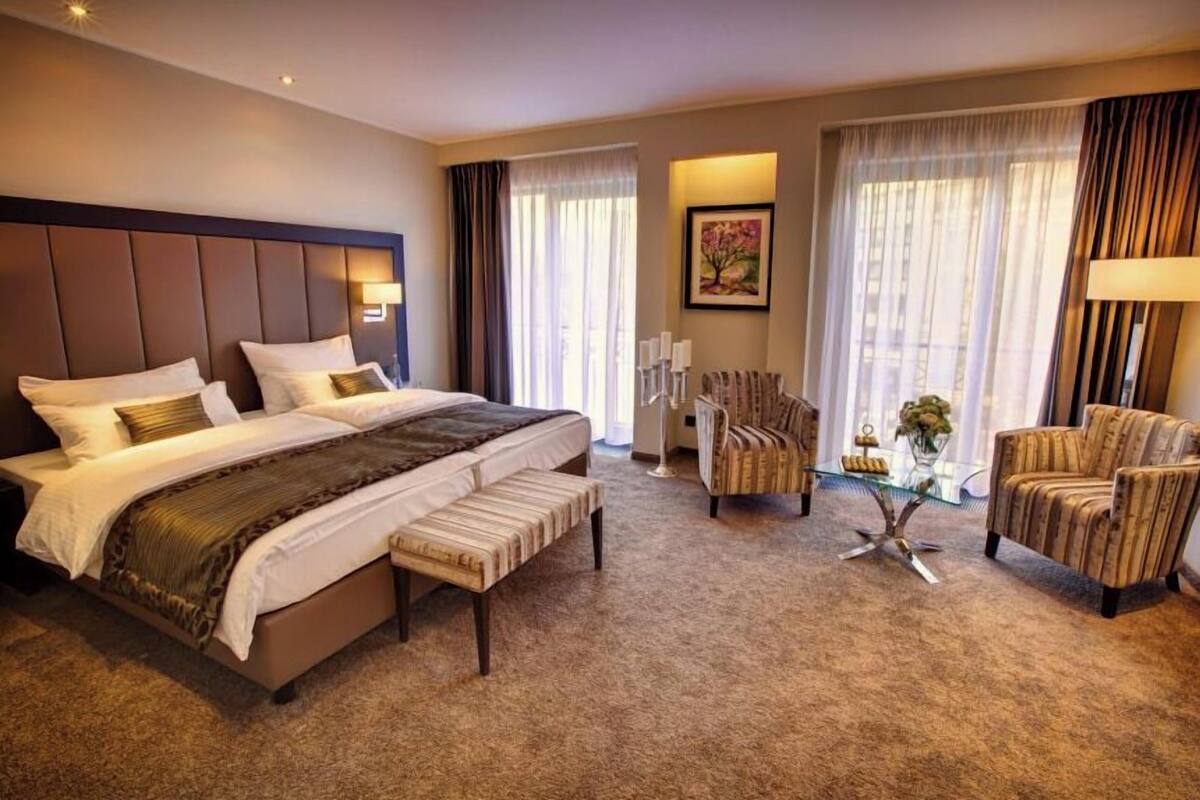 A hotel room with a large bed, a chair, a small table, and a painting on the wall.