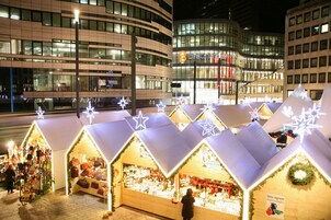 File di bancarelle luminose di un mercatino di Natale con decorazioni a stella, sullo sfondo di una facciata di un edificio moderno.
