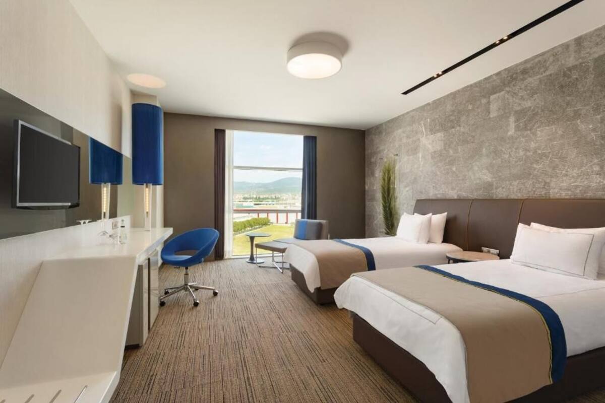 Ein Hotelzimmer mit zwei Betten, einem Schreibtisch, einem Sessel, einer Badewanne und einem großen Fenster mit Blick auf die Stadt.
