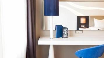 Habitación de hotel con un escritorio blanco, una silla azul, una lámpara y una cama con ropa de cama blanca.