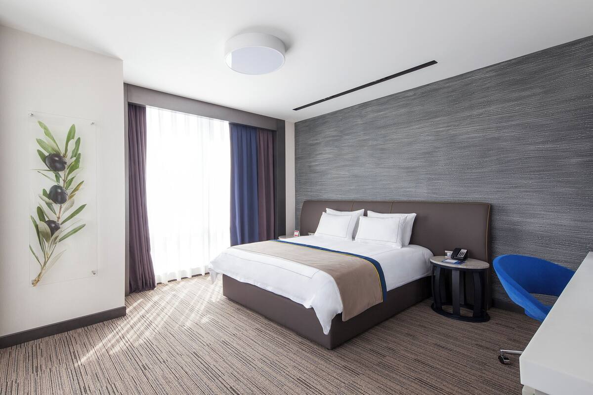 Ein modernes Hotelzimmer mit einem großen Bett, einem kleinen Beistelltisch, einem blauen Sessel und einem großen Fenster mit Vorhängen.