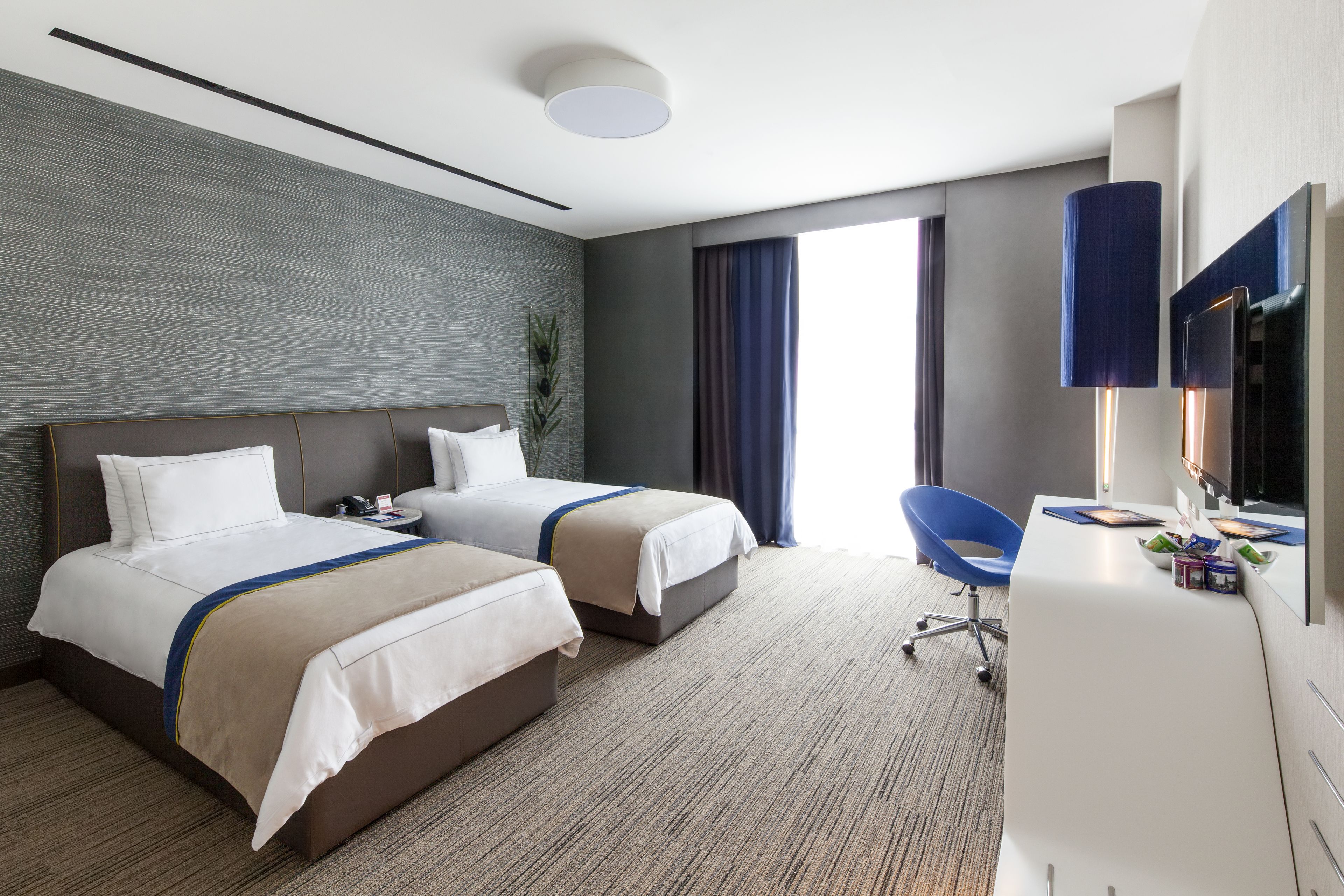 Habitación de hotel con dos camas, un escritorio, una silla, un televisor y un ventanal grande con cortinas.