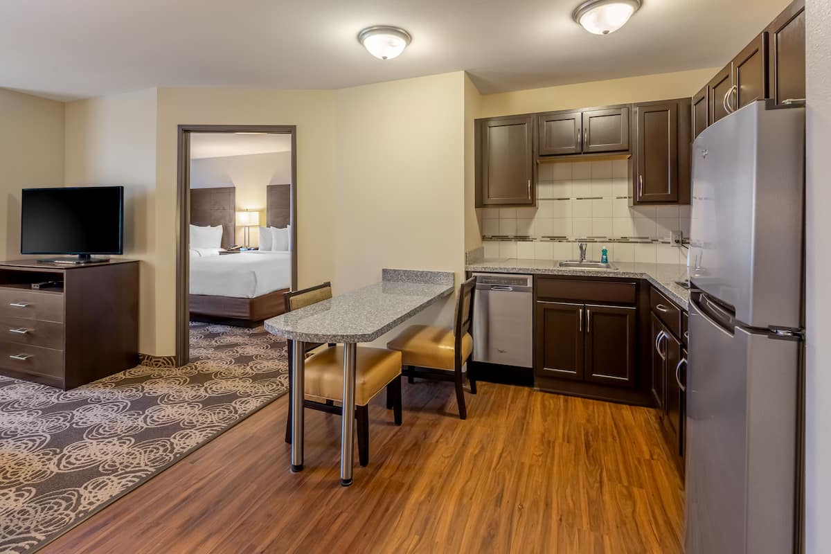 A compact hotel room with a kitchenette, a small dining area, a flat-screen TV, and a bed with a headboard.