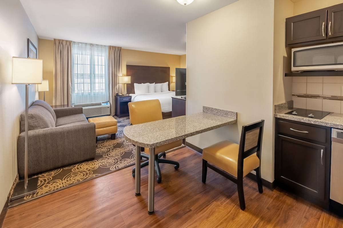 A hotel room with a bed, a desk, a chair, a sofa, a TV, a microwave, and a kitchen area.