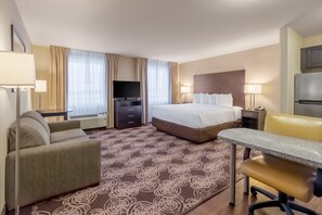 A hotel room with a bed, a desk, a chair, a television, and a refrigerator. - Landmark Suites (Williston)