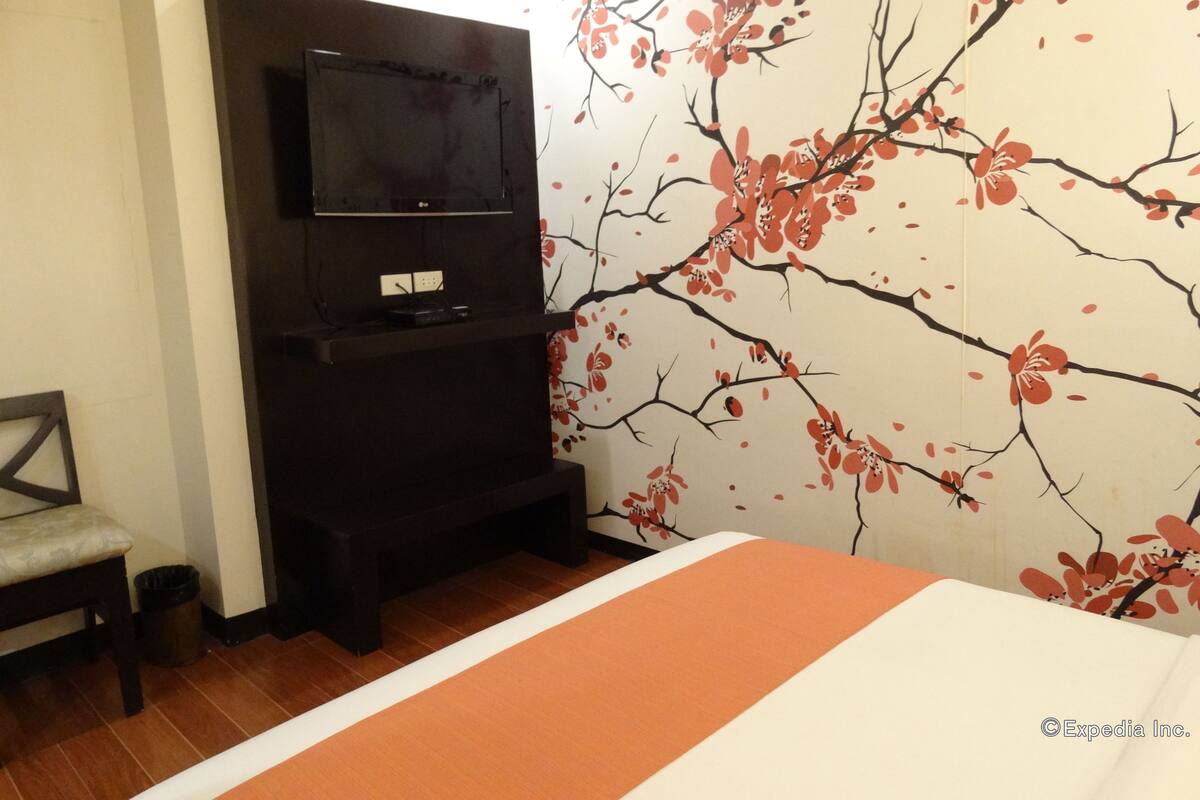 A hotel room with a bed, a TV mounted on a dark wooden stand, a chair, and a wall with a cherry blossom pattern.